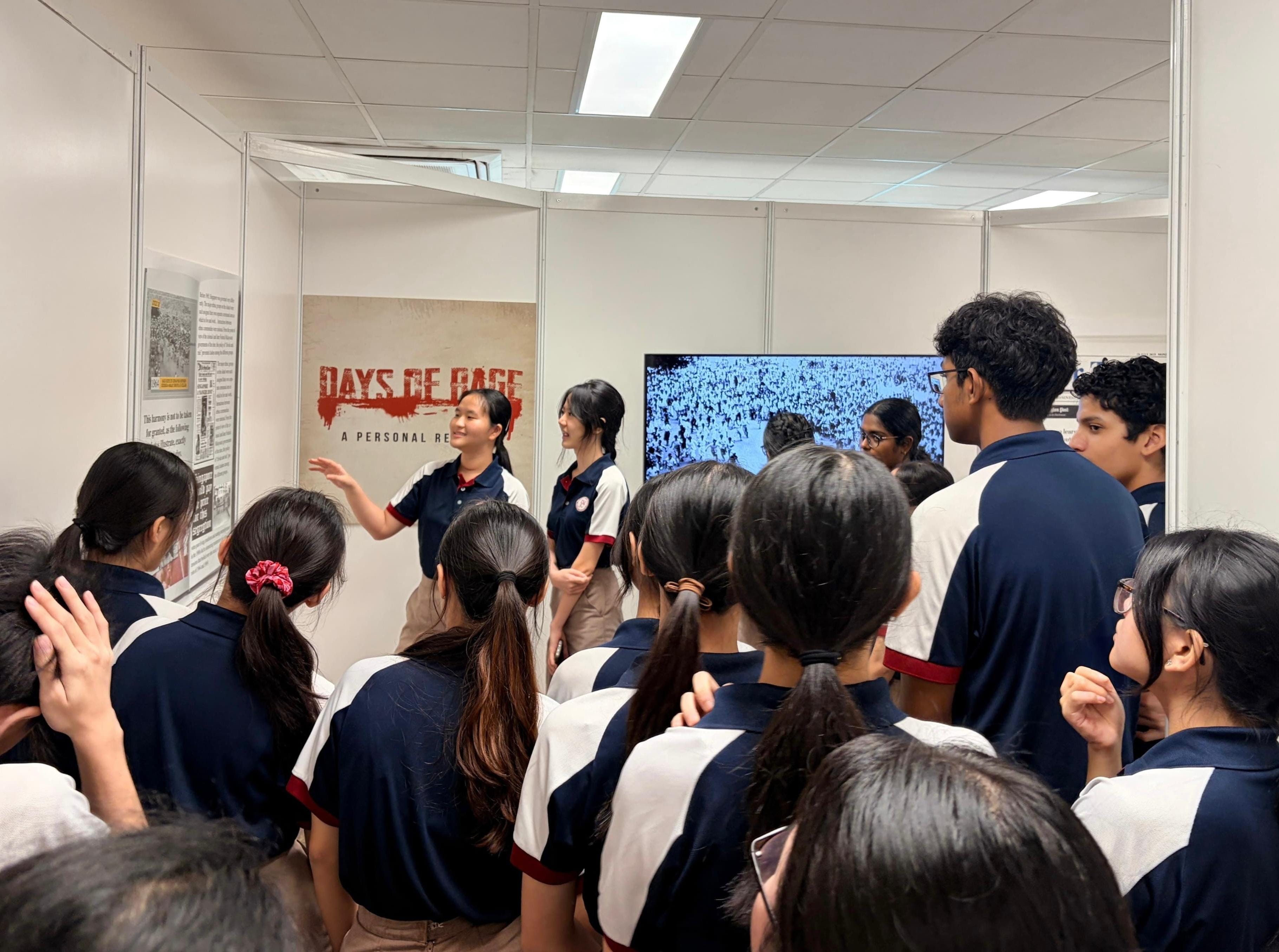 Two student leaders present to a group of classmates in front of a "Days of Rage" exhibition panel, with historical images displayed on a screen behind them.